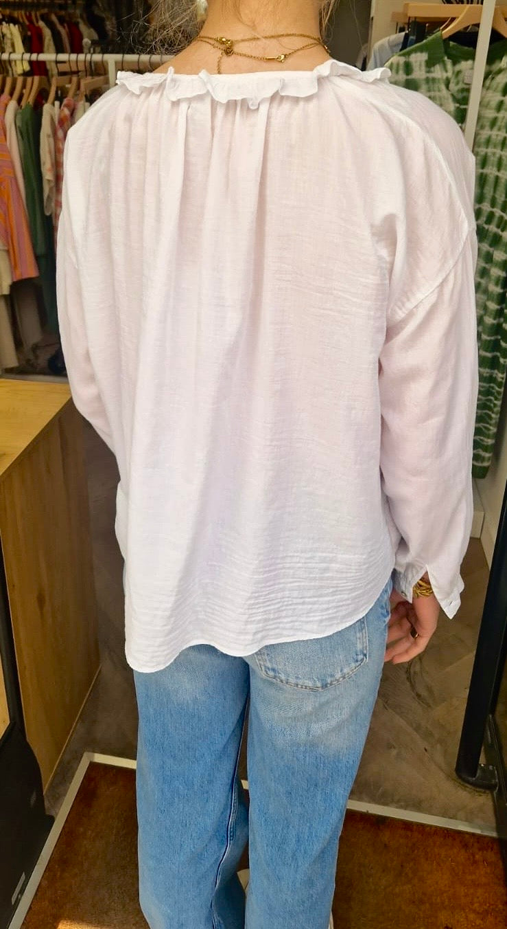 Vue de dos de la blouse blanche de chez Bella jones. Elle a un petit volant sur le col en V et des manches longues. Le dos est froncé.