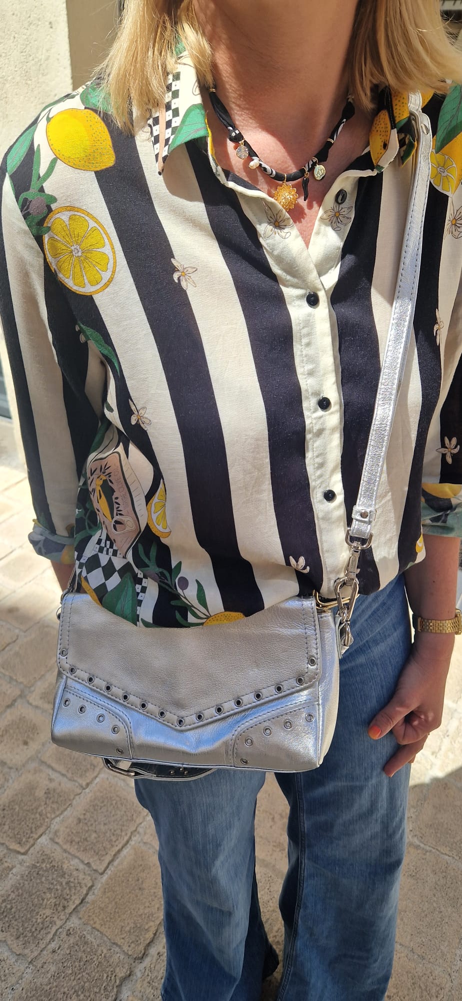 Sac Serena de la marque Neuville en cuir argenté porté en bandoulière. Vue en situation extérieure, avec détails cloutés et rabat structuré.
