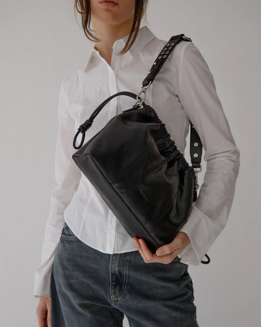 Sac Elliot Mini de la marque Neuville en cuir noir porté à l’épaule. Format compact, style souple et bandoulière cloutée.